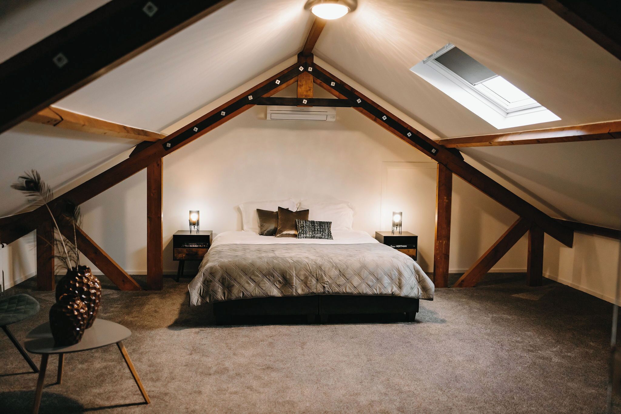 A bedroom with a sloped ceiling, a skylight, a bed with a quilted duvet, two bedside tables with lamps, and a small round table with a vase.