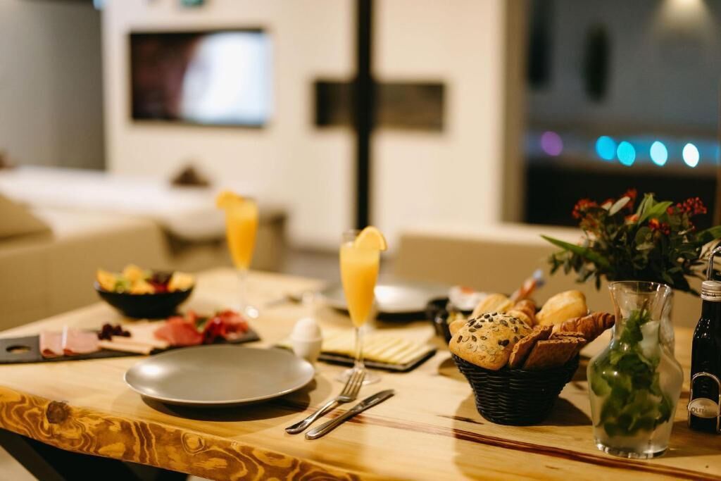 A breakfast setting with a table, a bowl of fruit, a glass of orange juice, bread, and a vase with flowers.