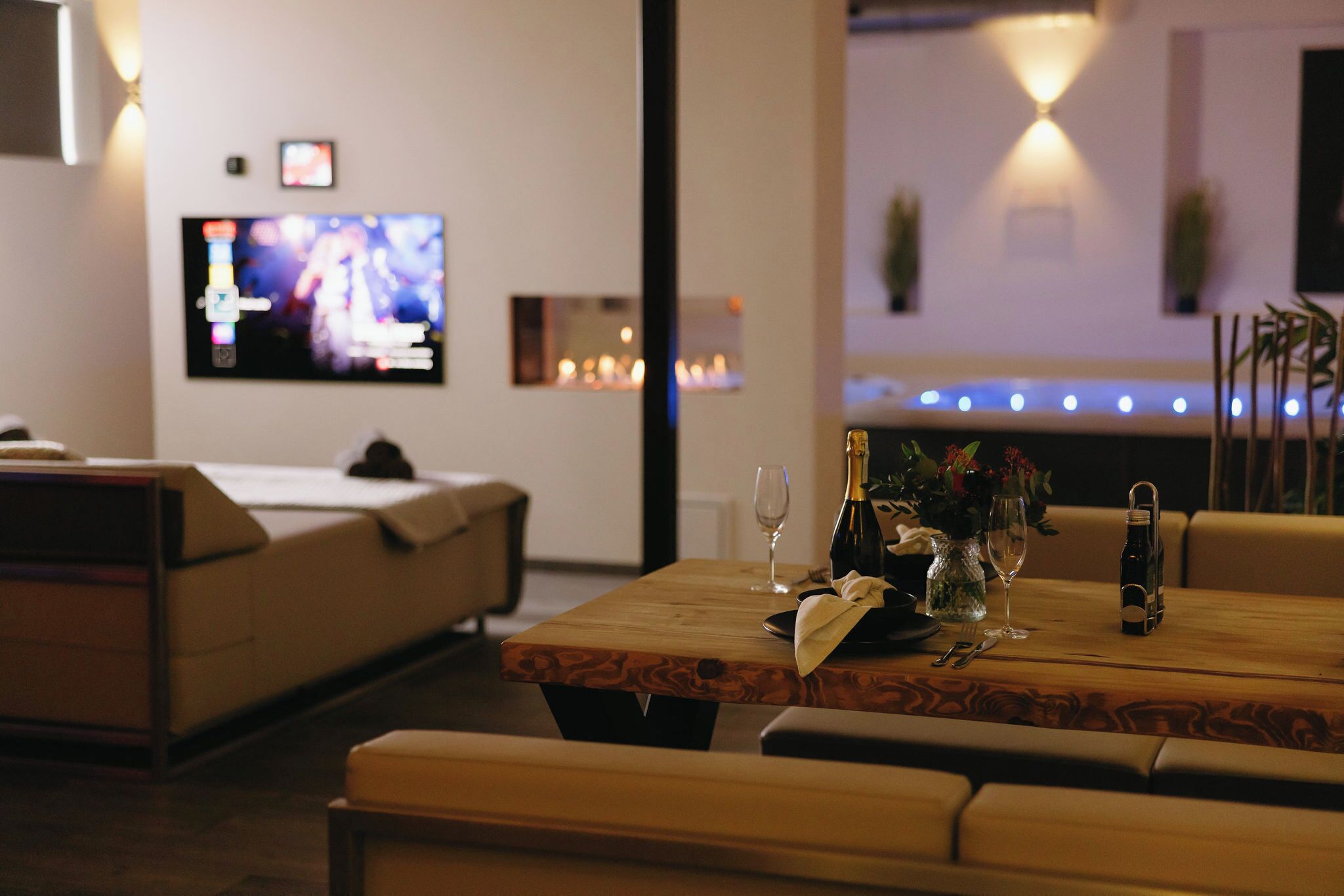 En moderne stue med en sofa, et træsofabord med en vase, vinglas og en vinflaske. Et fladskærms-tv er monteret på væggen, og der er dekorativ belysning i loftet.