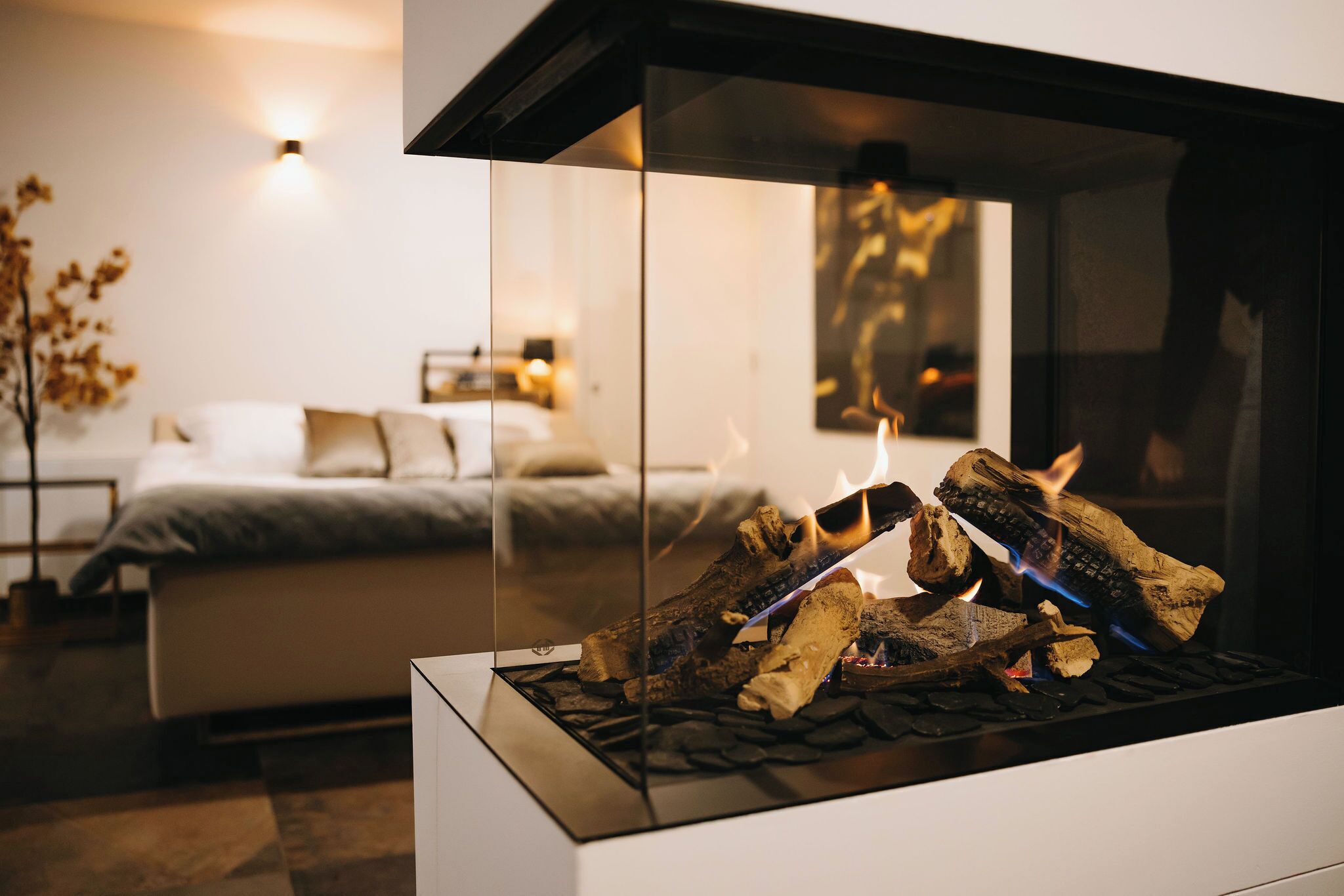 Ein modernes Wohnzimmer mit Kamin aus Glas, einem Bett in Grau und Weiß sowie einer Wandleuchte.