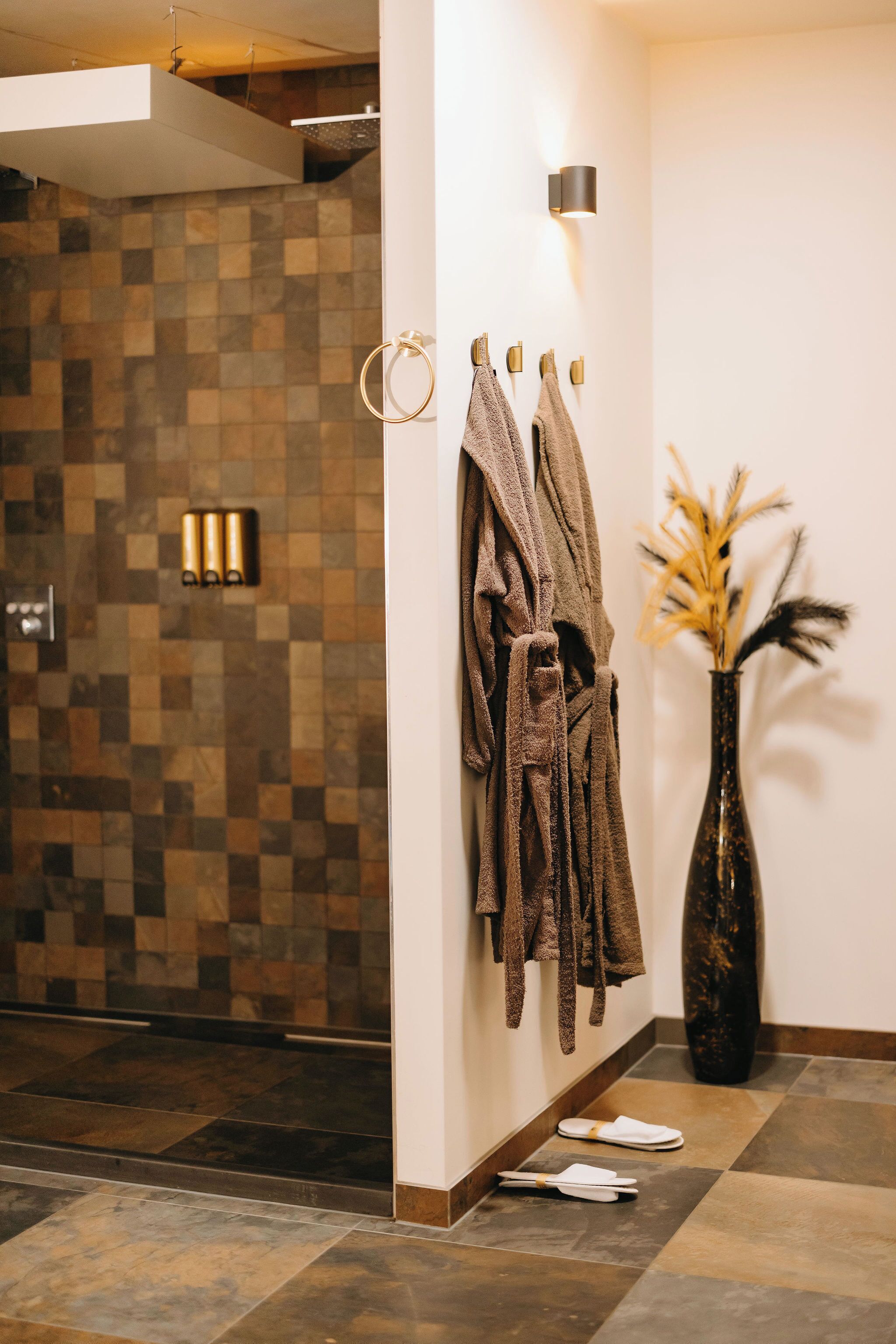 Un baño con pared de azulejos, un perchero con varias toallas, un florero con flores secas y un par de pantuflas en el suelo.