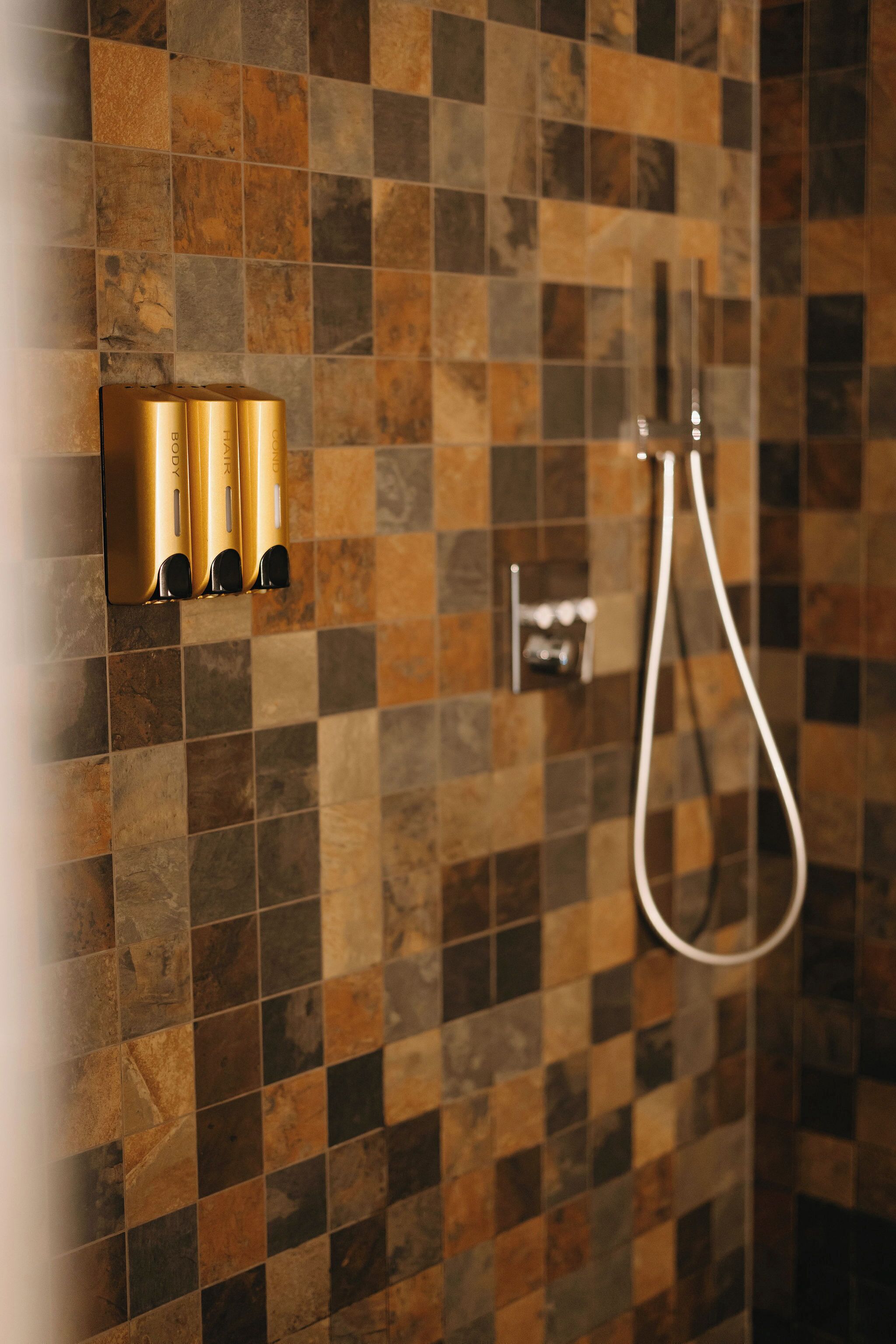A bathroom with a tiled wall, a shower, and a set of golden toiletries.