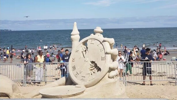 En strand med en stor sandskulptur föreställande en klocka, en stor folkmassa och havet i bakgrunden.