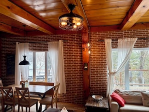 Ein gemütliches Zimmer mit Backsteinwand, Holzboden und einer Essecke mit Stühlen, einem Sofa und einem Fenster mit Vorhängen.