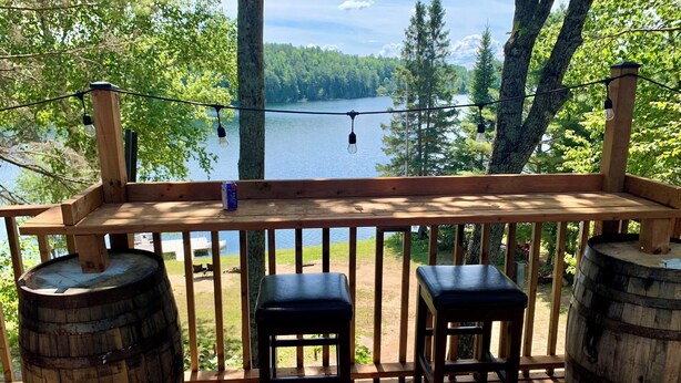 A wooden deck with two stools and two wooden barrels, overlooking a lake with trees and a clear sky. - The Hidden Gem (Gracefield)