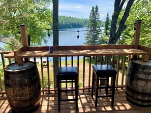 Une terrasse en bois avec deux tabourets et deux tonneaux en bois, donnant sur un lac bordé d’arbres et sous un ciel dégagé.