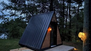 A small, triangular wooden cabin with a sloped roof, a door, and a small porch area with lights. - The Hidden Gem (Gracefield)