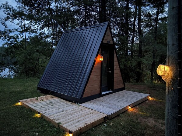 Une petite cabane triangulaire en bois, dotée d’un toit en pente, d’une porte et d’un petit porche éclairé.