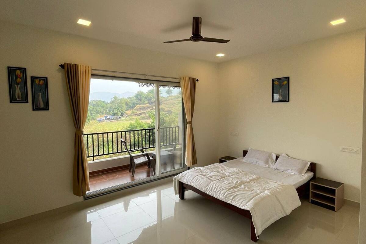 A bedroom with a bed, a ceiling fan, a balcony with a view of greenery, and a window with curtains.