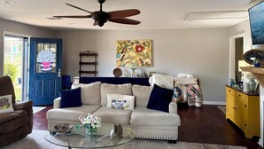 Un salon avec un ventilateur de plafond, une porte bleue, un canapé beige, une table basse en verre et un meuble jaune.
