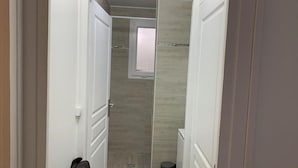 Un couloir étroit, avec une porte sur la gauche, une fenêtre au fond et une petite poubelle sur la droite.