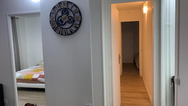 Un couloir avec une horloge au mur, un lit recouvert d’une couverture à rayures et une porte donnant sur une autre pièce.