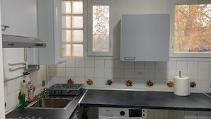 Une cuisine dotée d’un évier en acier inoxydable, d’un égouttoir, d’une fenêtre avec stores et d’un mur blanc orné de carreaux à motifs floraux.