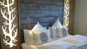 A neatly made bed with white bedding and decorative pillows, a wooden headboard with a tree light design, and a bedside table with a phone charger. - Landhotel Talblick  (Neuweiler)