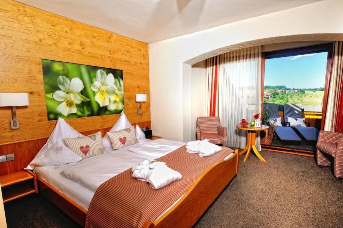 A hotel room with a wooden headboard, a large bed with white bedding, a painting of flowers, a balcony with a view, and a small table with a chair.