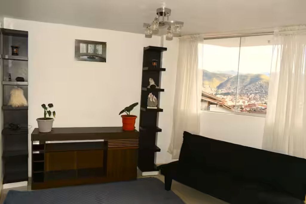 Ett rum med en säng, en svart soffa, ett skrivbord med en växt, en hylla med dekorativa föremål och ett fönster med utsikt över staden.