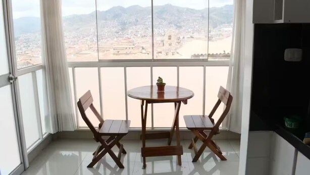 Ein Balkon mit Tisch und zwei Stühlen, einem Blumentopf und Blick auf eine Stadtlandschaft mit Bergen.