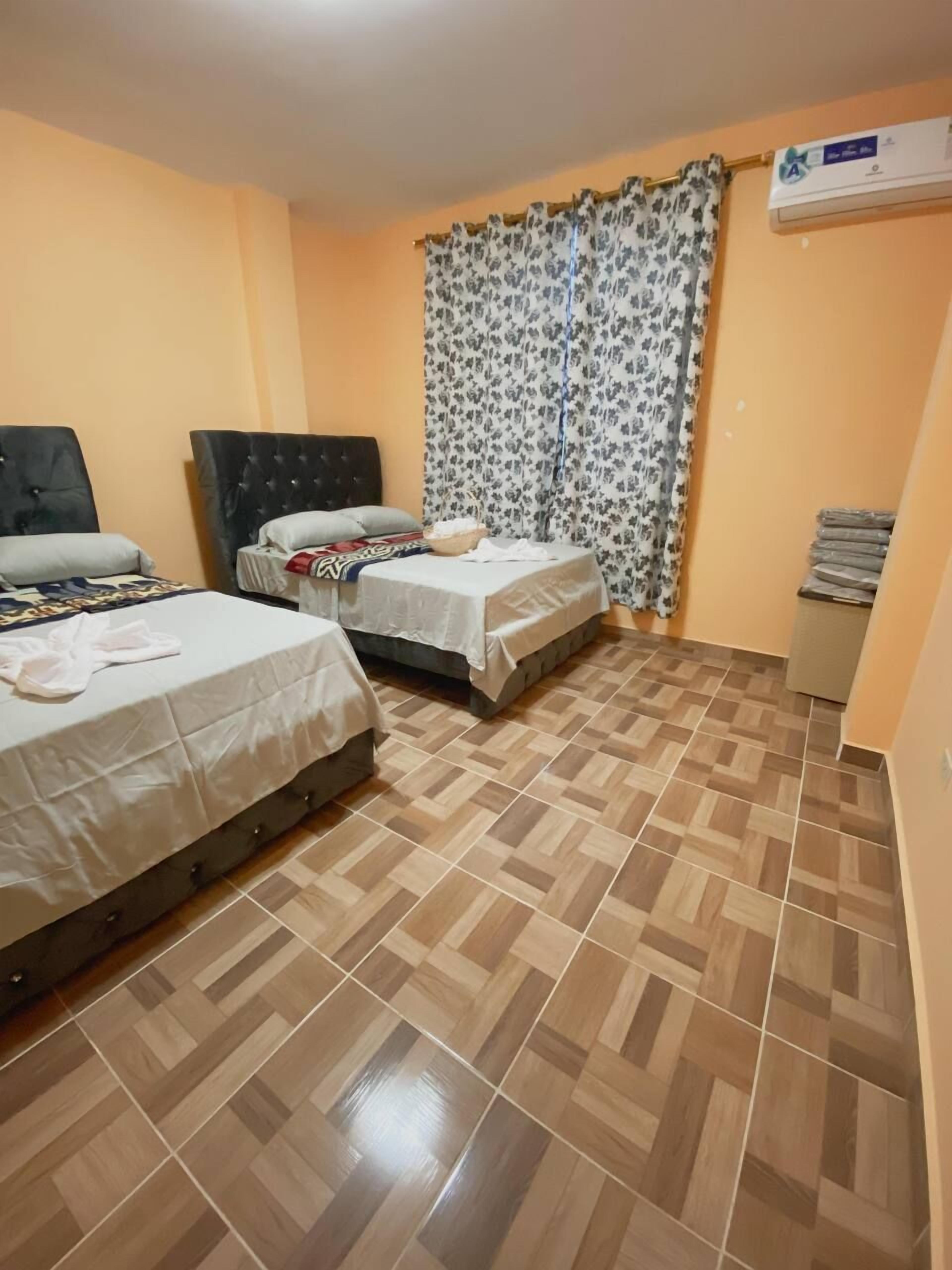 A room with two beds, a window with floral curtains, a tiled floor, and an air conditioning unit.