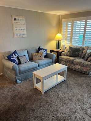 Ein Wohnzimmer mit grauem Sofa, einem weißen Couchtisch und einer Wand mit einem Schild im Strandhaus-Stil.