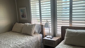 Une chambre avec deux lits, une table de chevet, une lampe et une fenêtre avec des stores.