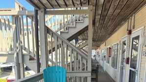 Un espace extérieur abrité avec des chaises turquoise et une table en bois, un escalier en bois et un bâtiment doté de grandes fenêtres et d’une porte.
