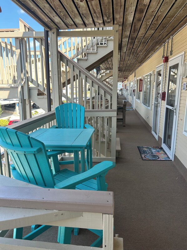 A covered outdoor seating area with turquoise chairs and a wooden table, a wooden staircase, and a building with large windows and a door. - Cozy Condo in Ocean City steps from the beach (Ocean City)