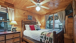 Une chambre à coucher dans une cabane en bois, comprenant un lit, une table de chevet, une commode, un ventilateur de plafond et de grandes fenêtres.