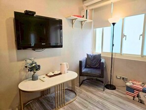 A living room with a flat-screen TV mounted on the wall, a small table with a vase and a kettle, a grey armchair, a floor lamp, and a window with blinds. - [2026/4 NEW RENOVATION] Bright Large Window Ensuite Quiet Cozy Room-1 Min to MRT (Taipei City)