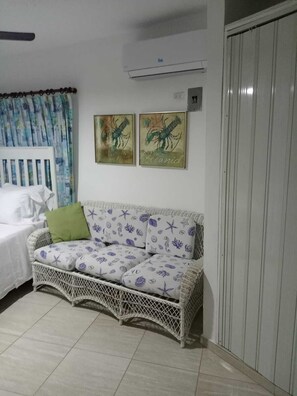 Une pièce avec un canapé en osier blanc, un lit avec une literie blanche et deux tableaux accrochés au mur.