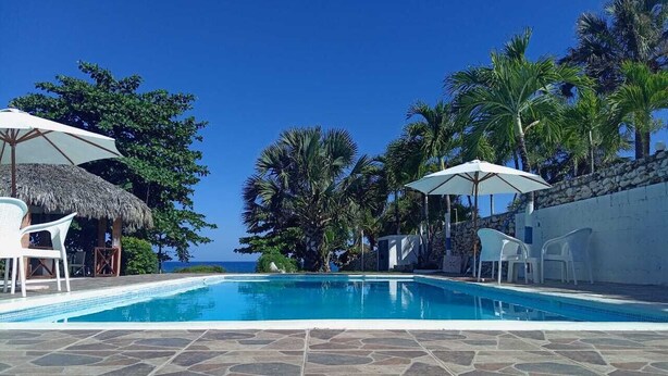Una piscina con baldosas de piedra, sillas de descanso blancas y sombrillas, rodeada de palmeras y un cielo despejado.