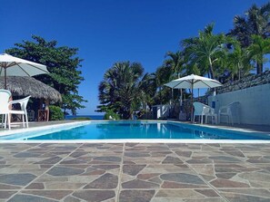Une piscine aux carreaux de pierre, des chaises longues blanches et des parasols, le tout entouré de palmiers et surplombé par un ciel dégagé.