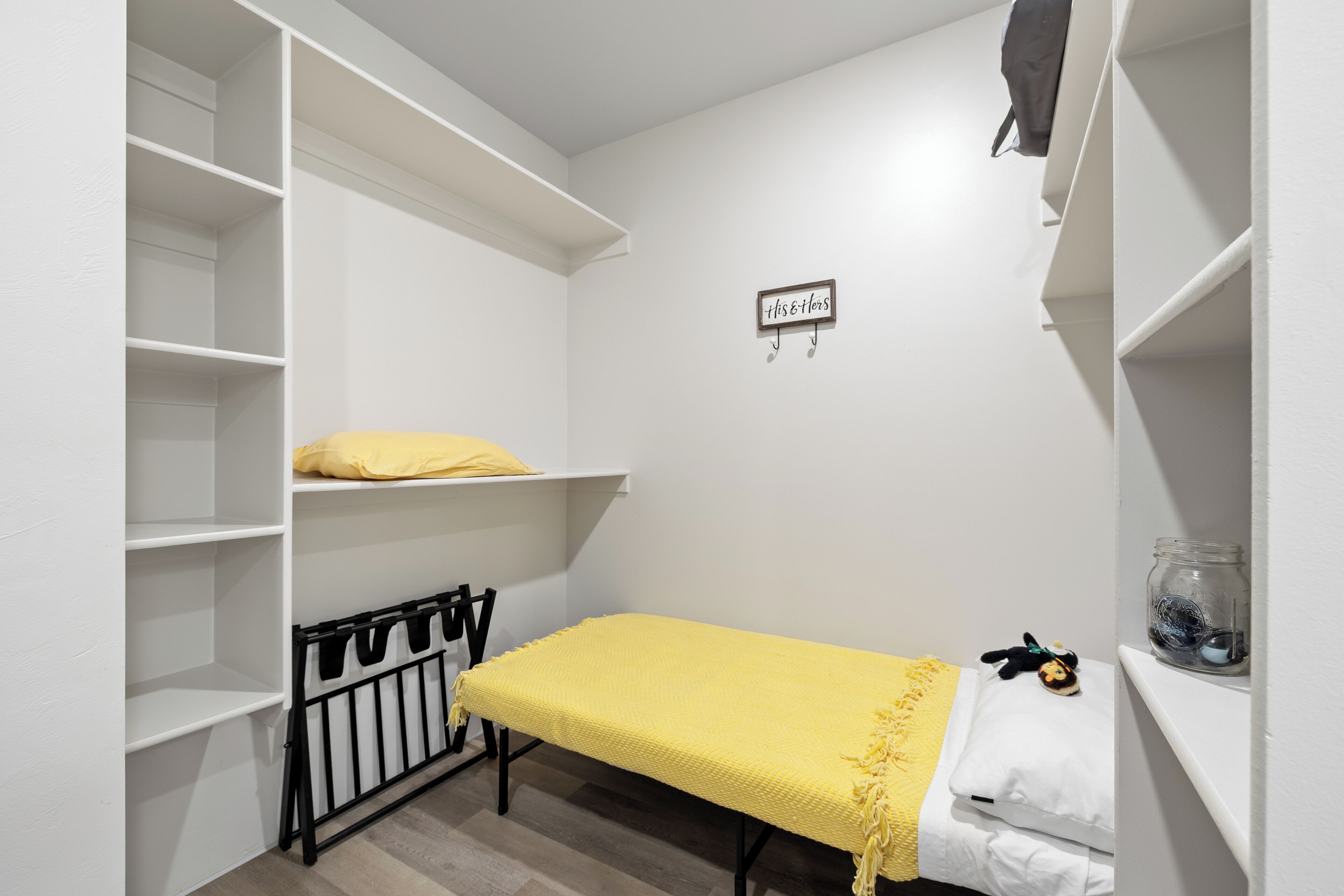 A small, neatly organized room with a single bed, a shelf with a yellow pillow, a wall-mounted sign, and a black metal crib.