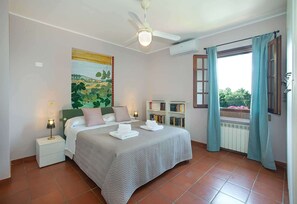 Ein Schlafzimmer mit einem Bett, Nachttischen, einem Deckenventilator, einem Gemälde, einem Bücherregal und einem Fenster mit Vorhängen.