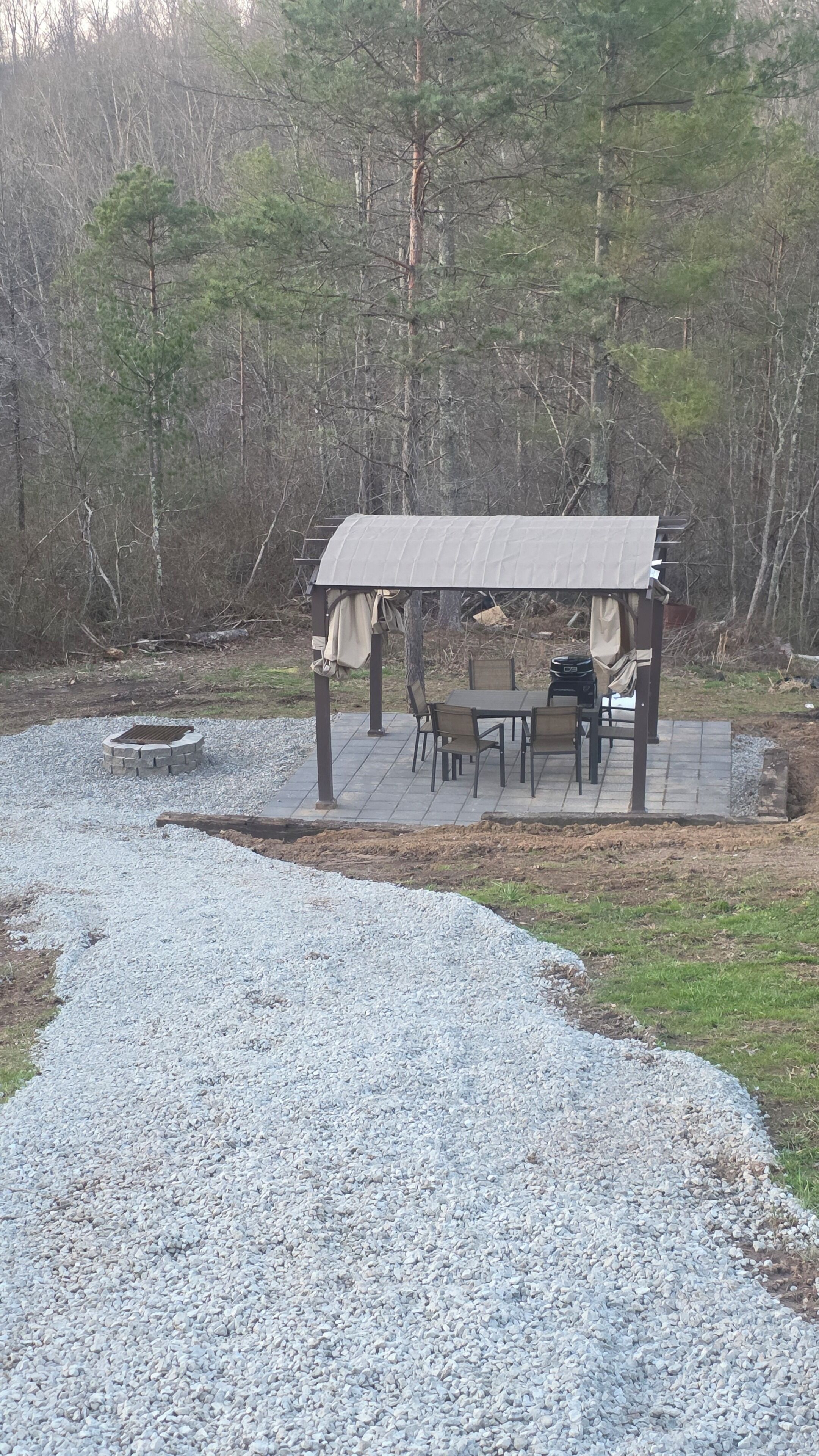 Een overdekt terras met een tafel en stoelen, omgeven door een grindpad en bebossing.