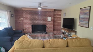 Una sala de estar con chimenea de ladrillo, ventilador de techo, televisor sobre un mueble de madera y sofá.