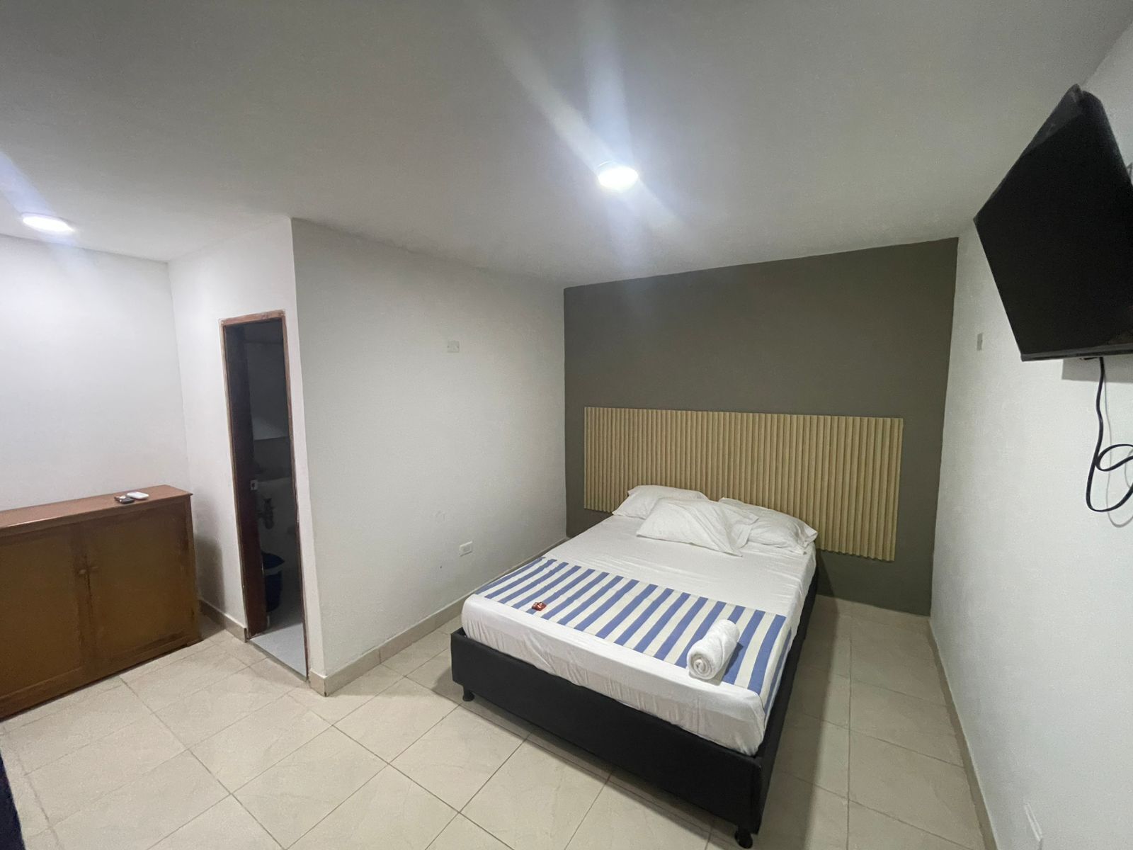 Un dormitorio con cama, un escritorio de madera, televisor montado en la pared y una puerta que da a otra habitación.