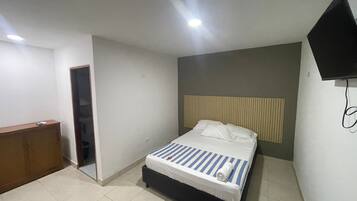 Un dormitorio con cama, un escritorio de madera, televisor montado en la pared y una puerta que da a otra habitación.