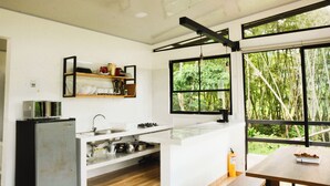 Uma cozinha com pia, fogão e prateleiras, grandes janelas com vista para o verde, e uma pequena mesa com um prato.