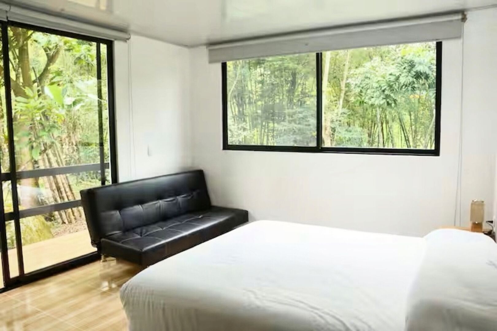 Un dormitorio con cama, sofá de cuero negro, ventanas amplias y vistas a zonas verdes.