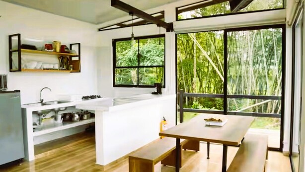 Cozinha moderna com ilha central, piso de madeira e grandes janelas com vista para uma floresta.