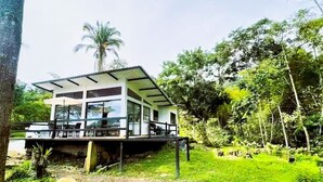 Uma casa moderna de dois andares com varanda, cercada por vegetação exuberante e uma palmeira.