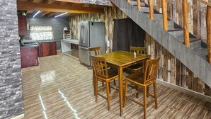 Ruime kamer met een houten trap, een eetgedeelte met tafel en stoelen, en een keuken met een koelkast en kastjes.
