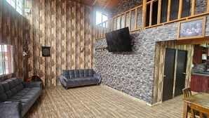 Een ruime woonkamer met een hoge plafond, houten balken, een stenen muur en een wandgemonteerde televisie.