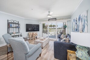 A living room with a TV, a ceiling fan, a bookshelf, and a view through a sliding glass door. - Renovated Spacious Condo! Two Resort Pools! (Miramar Beach)