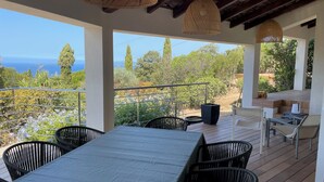 Ein überdachter Essbereich im Freien mit Meerblick, ausgestattet mit Tisch, Stühlen, einem Grill und hängenden Leuchten.