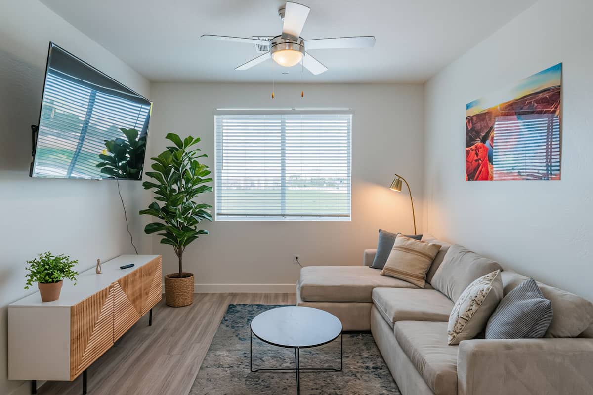 A living room with a sofa, a coffee table, a TV, a ceiling fan, a large window, and a plant.