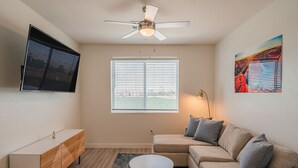 A living room with a ceiling fan, a TV mounted on the wall, a sofa with cushions, a small round coffee table, and a window with blinds. - Sleek Stay 4mins to State Farm Stadium (Glendale)