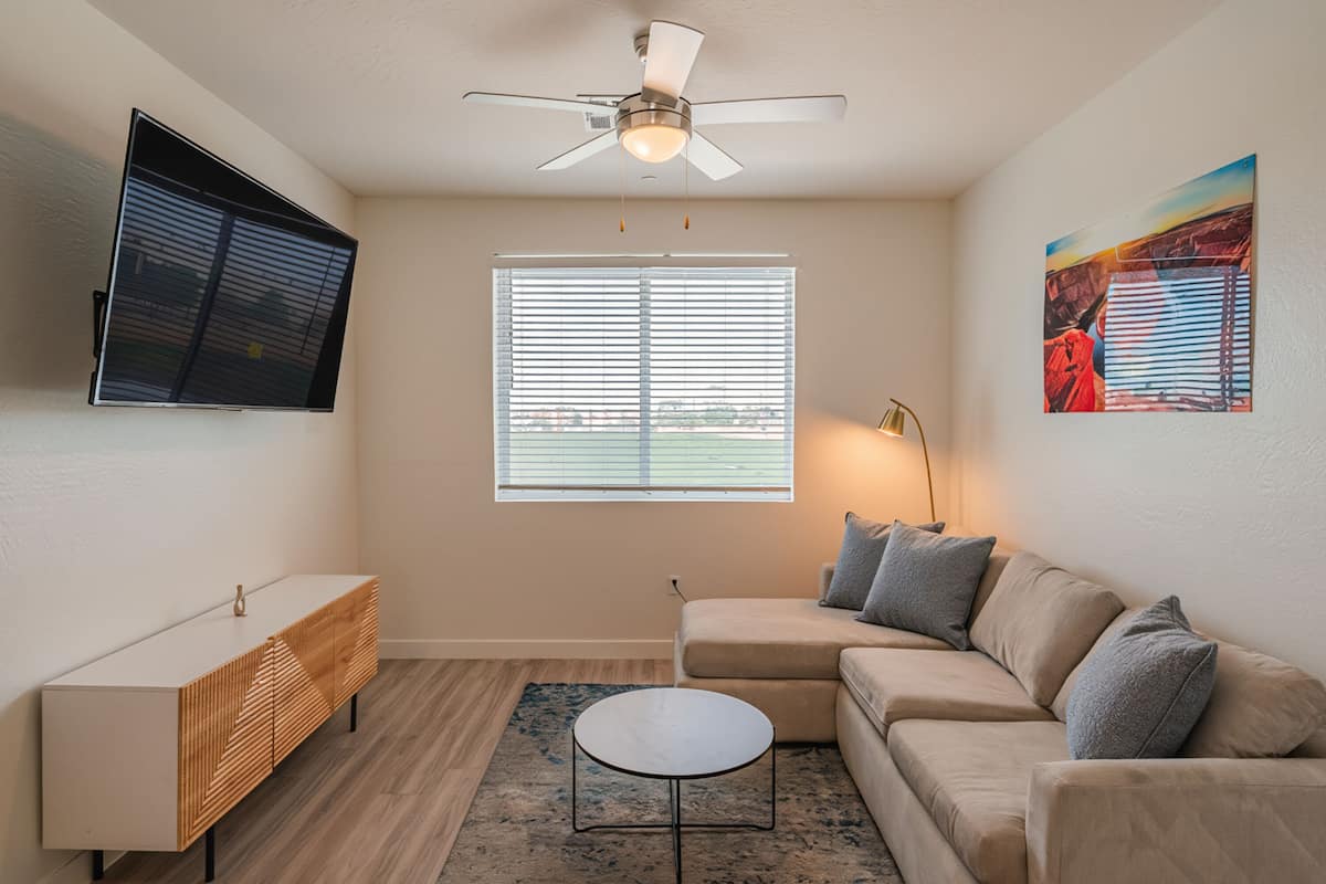 A living room with a ceiling fan, a TV mounted on the wall, a sofa with cushions, a small round coffee table, and a window with blinds.