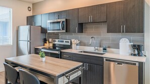 A modern kitchen with dark cabinetry, stainless steel appliances, a wooden dining table, and a dishwasher. - Sleek Stay 4mins to State Farm Stadium (Glendale)