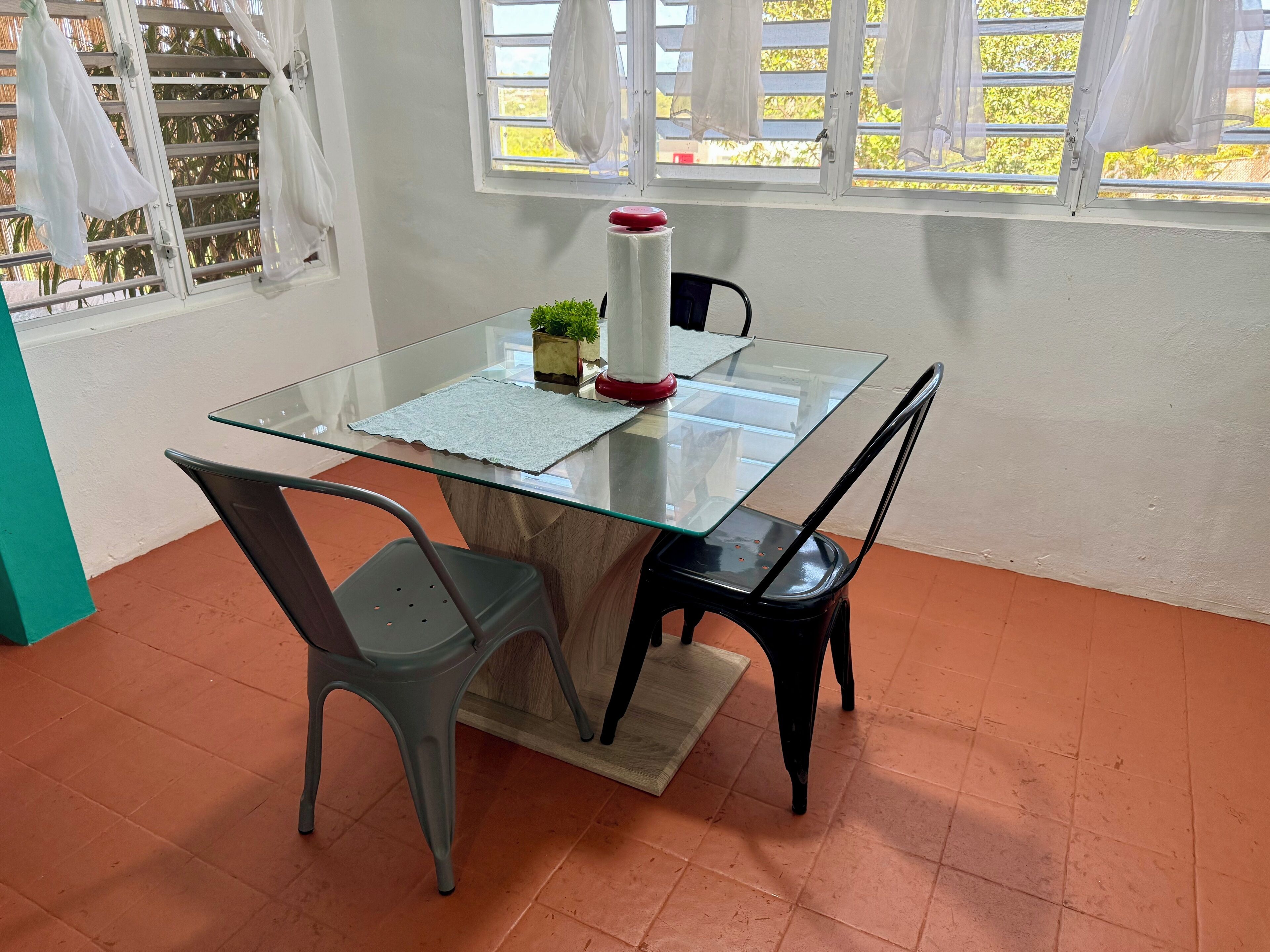 A glass-top table with a placemat, two chairs, a small plant, and a red object on the table.